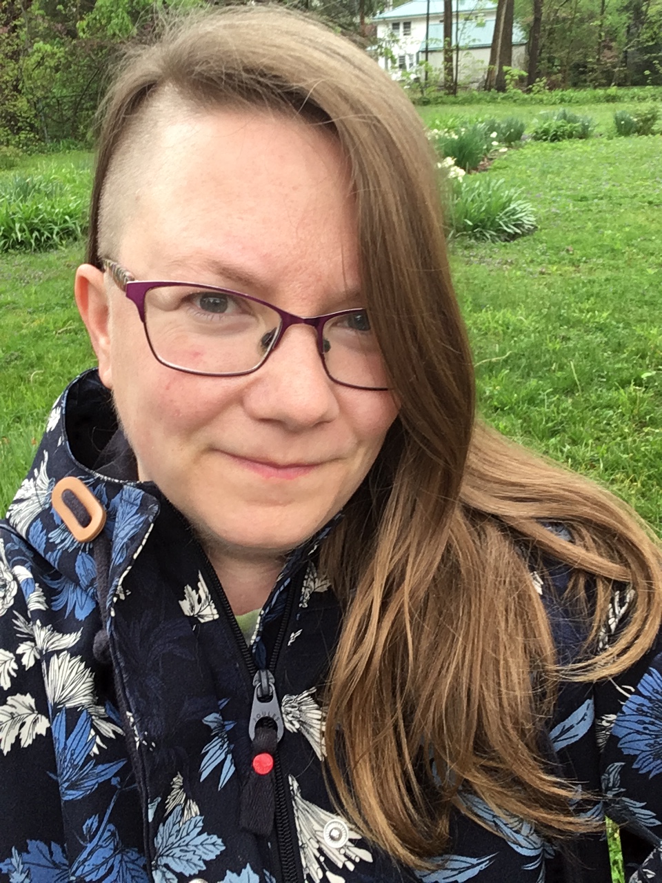 Photo of blog author, River, wearing a blue raincoat. They are wearing glasses, and have long hair, and a close-side shave. Behind them is green grass and flowers.