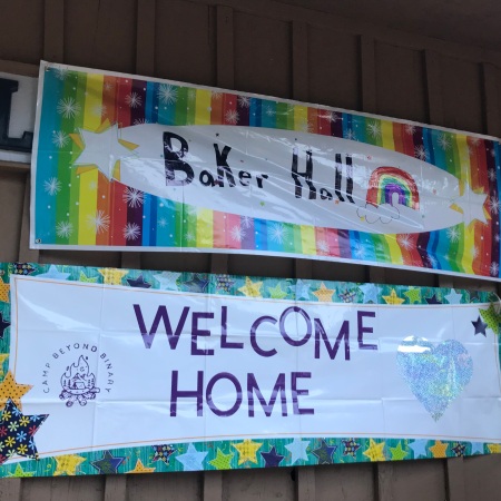 A rainbow banner that says "Baker Hall" with another banner below that says "Welcome Home." On the right side of the frame is a photo of Gilbert Baker who made the first Pride flag.